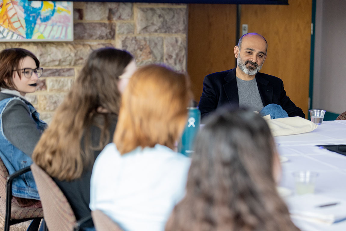 A bald man with a beard sits at a table with a row of college students sitting near him